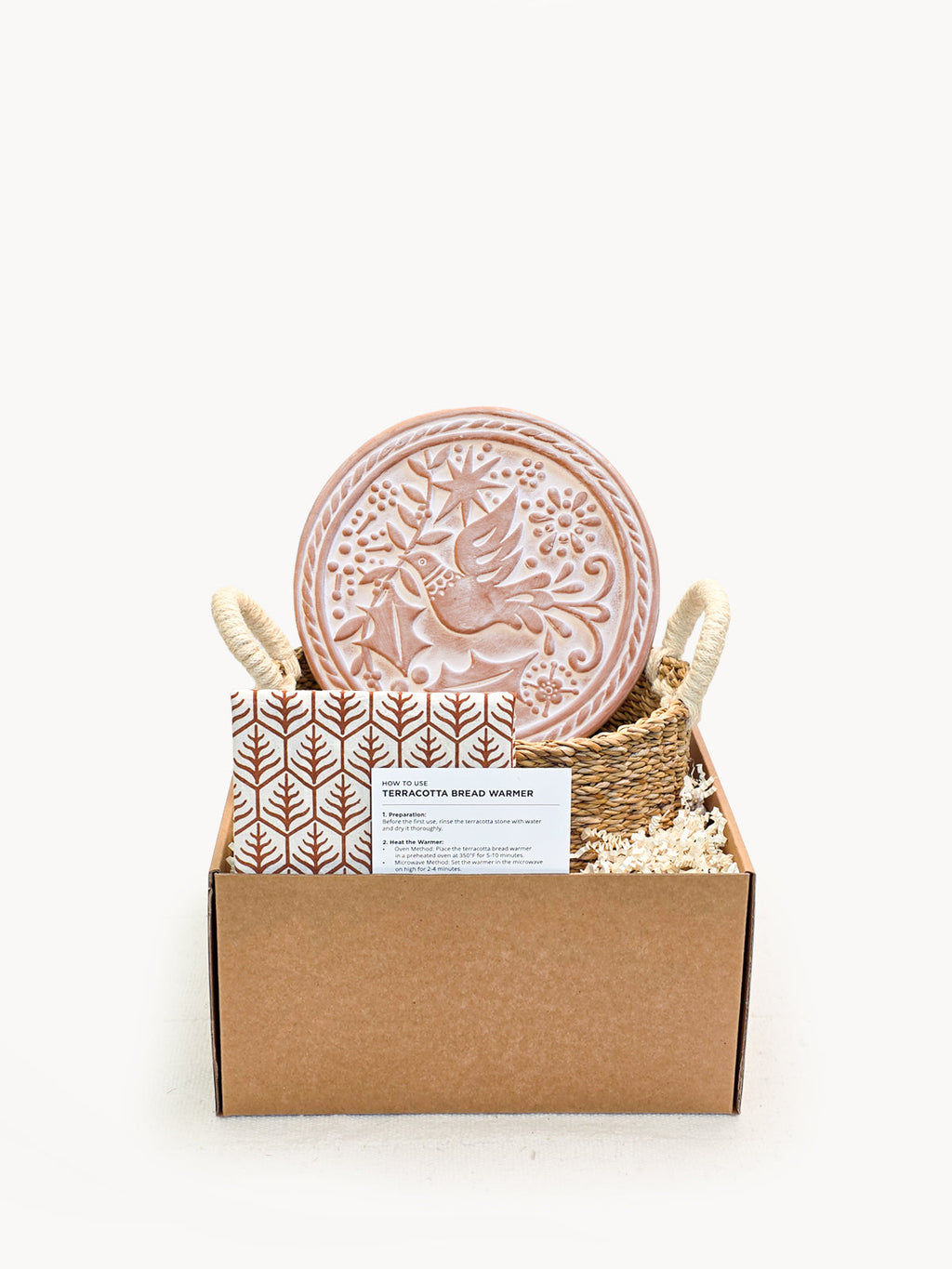 Round terracotta bread warmer with bird engraving inside a natural seagrass basket and cotton tea towel