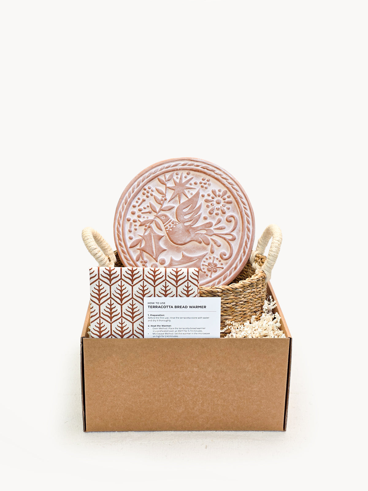 Round terracotta bread warmer with bird engraving inside a natural seagrass basket and cotton tea towel