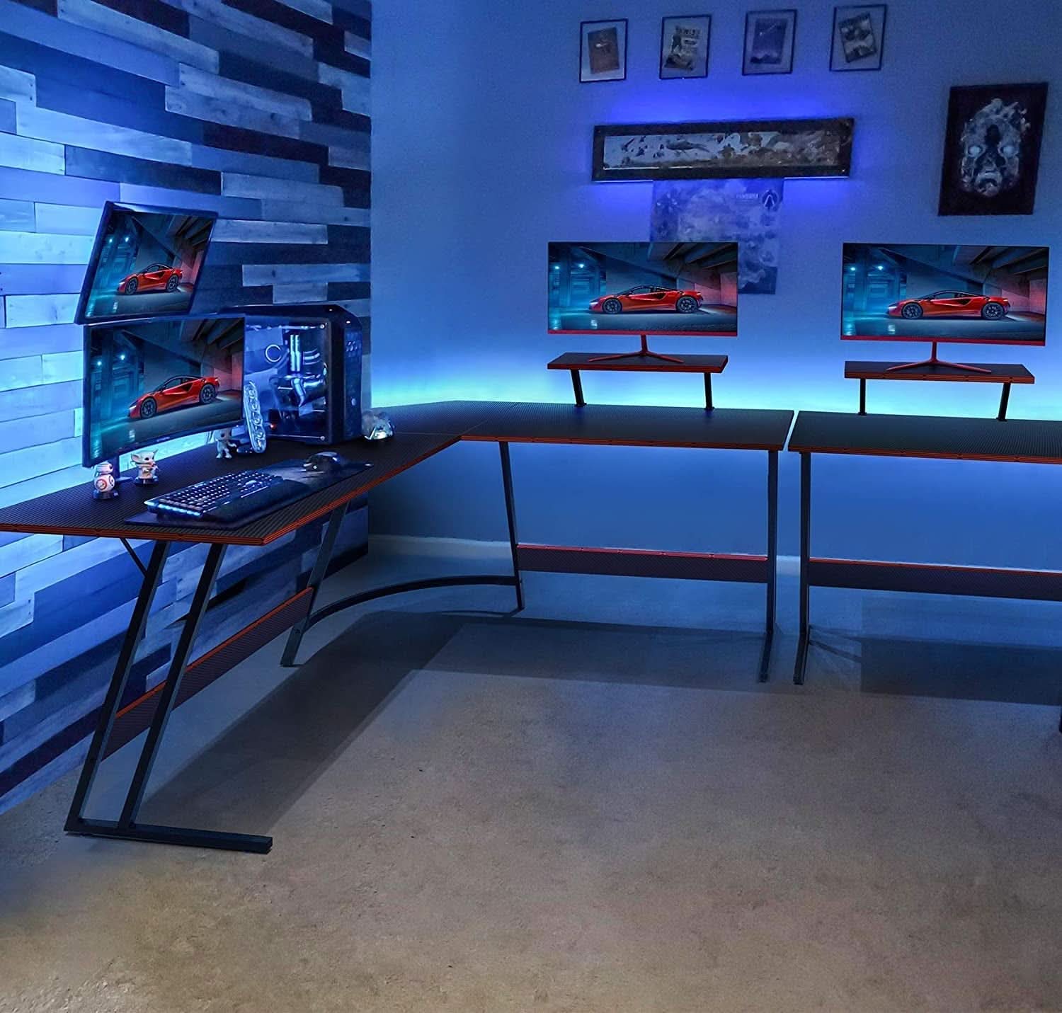 Black L-shaped gaming desk with carbon fiber finish, red accents, monitor riser, and metal Z-shaped legs — view 2