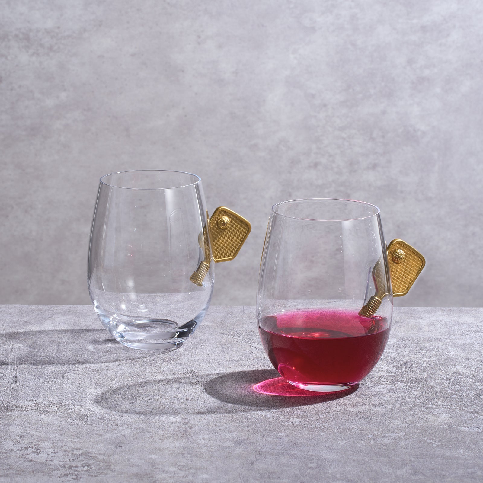 Two pickleball stemless wine glasses with decorative 3D gold racket details on a plain background — view 2