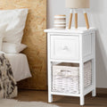 Two rustic white wood bedside tables with smooth top drawers and wicker storage baskets.