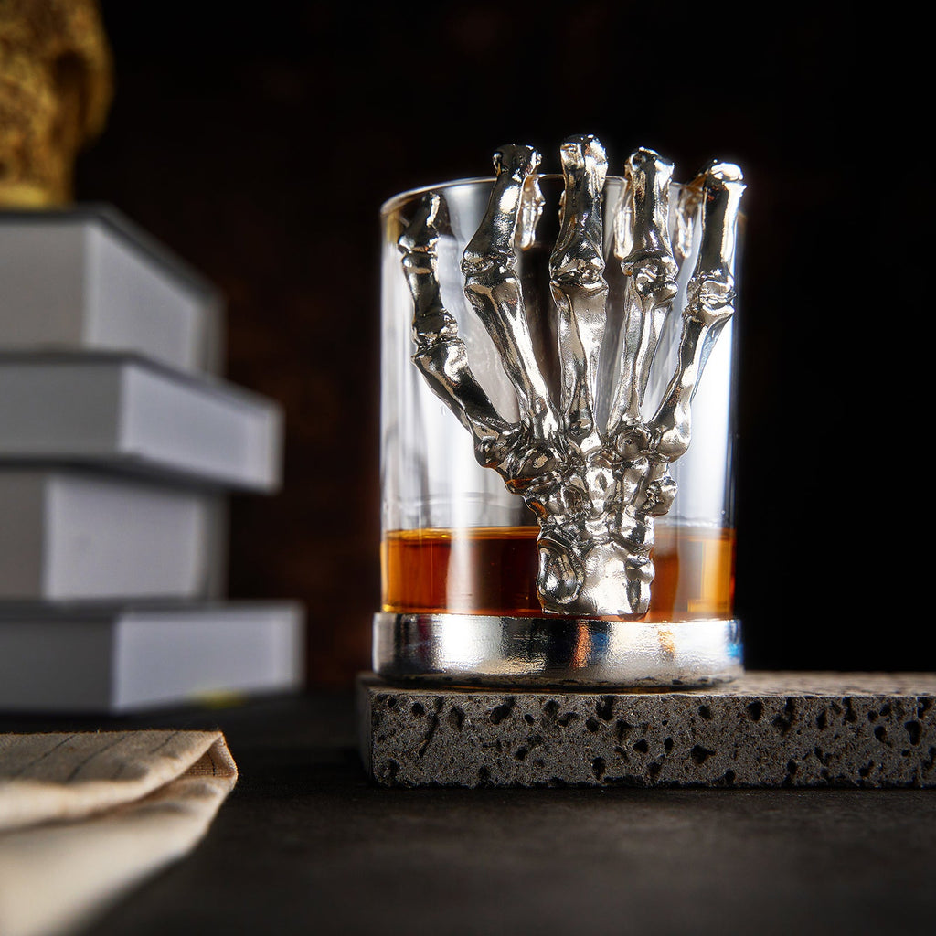 Skeleton whiskey glass tumbler with detailed bone hand stem, clear glass cup holding amber liquor — view 2