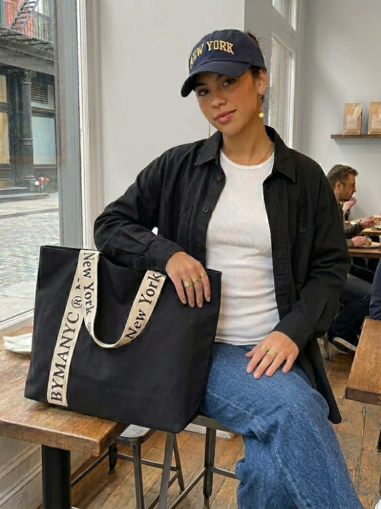 Women's black canvas tote bag with wide cream straps and bold typographic logo lettering.