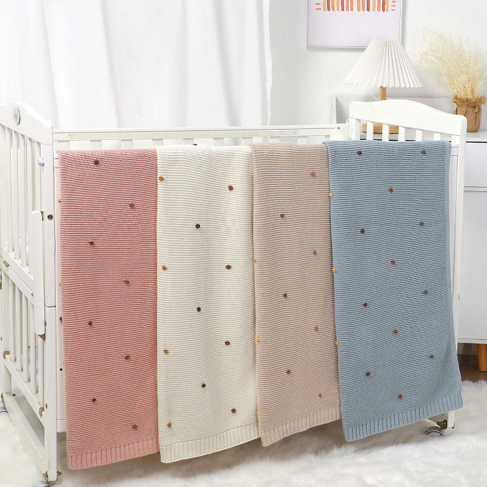 Knitted cotton baby blanket with hand-embroidered pom poms, in soft solid colors for contemporary nurseries.